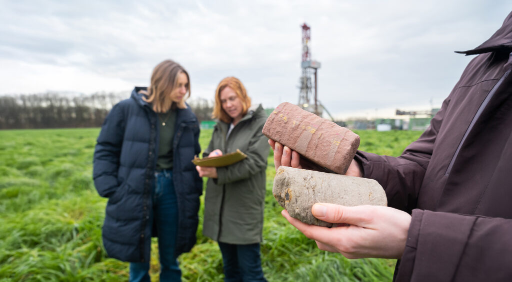 Handen houden twee boorkernen van steen vast op een grasveld, met een boortoren en twee personen op de achtergrond.