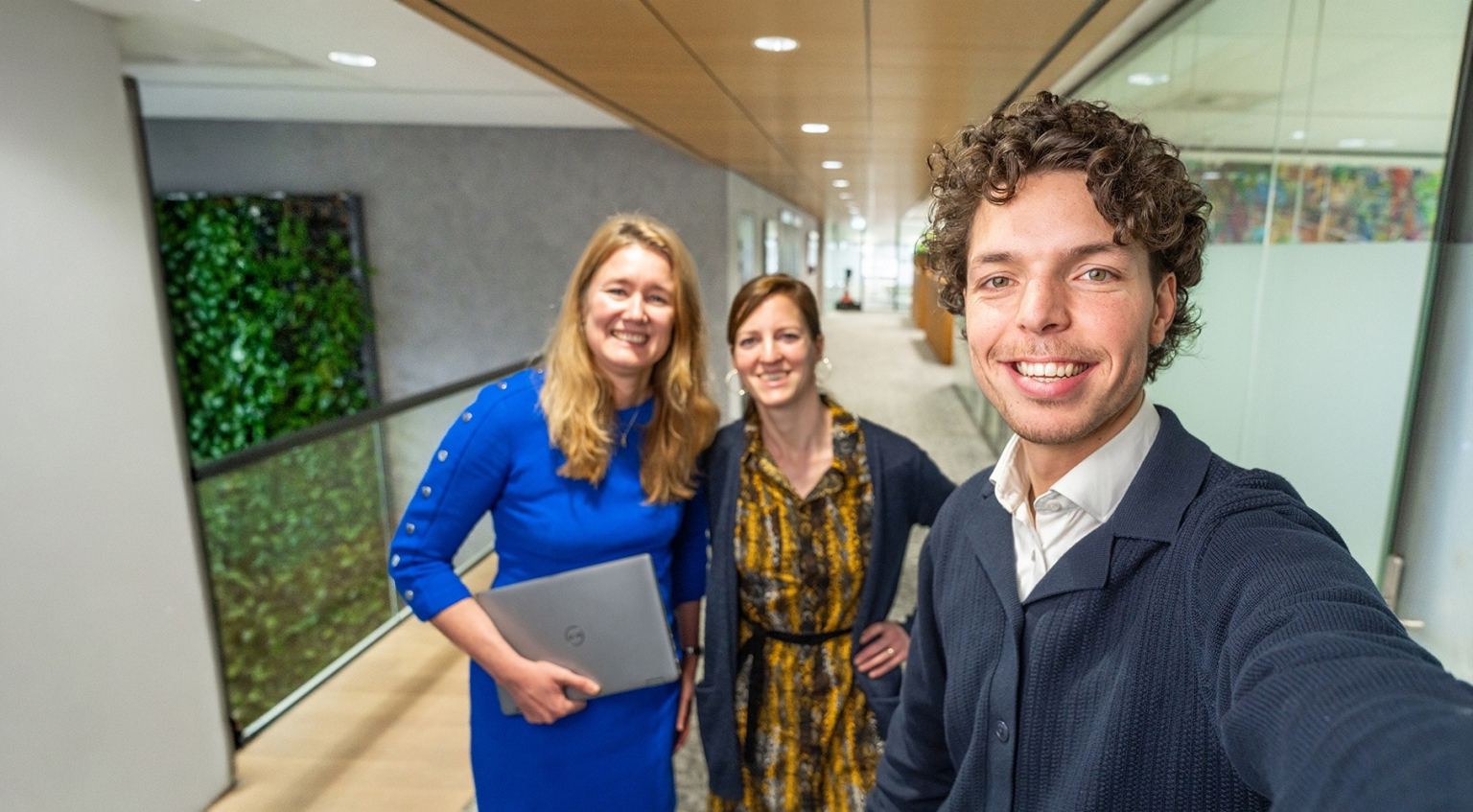 Drie EBN-medewerkers maken een selfie in een gang van het EBN-kantoor. Het kantoor is modern en licht met glazen wanden en een groene plantwand.