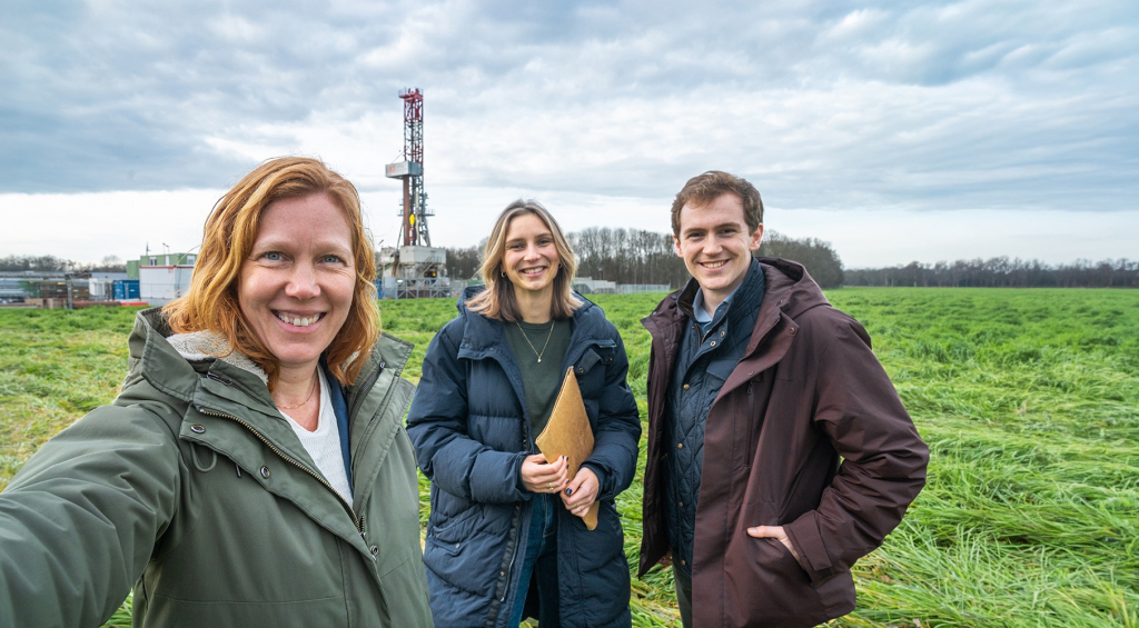 Drie EBN-medewerkers staan op een grasveld met een boortoren op de achtergrond. De boortoren voert onderzoek uit naar aardwarmte.