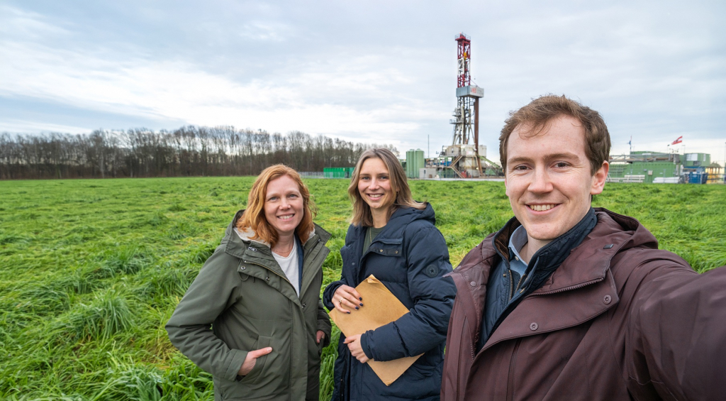 Drie EBN-medewerkers staan op een weiland met een boortoren en installaties op de achtergrond.
