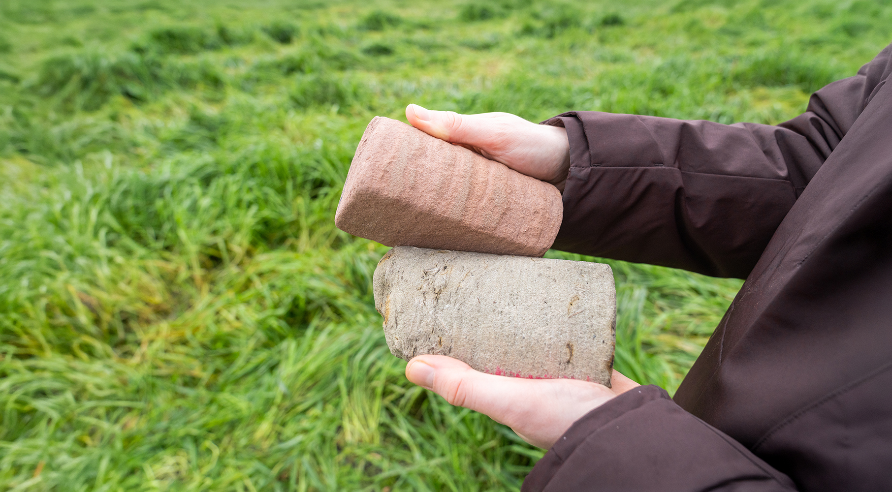 Handen houden twee boorkernen vast op een grasveld. Deze boorkernen zijn met geologisch onderzoek naar aardwarmte naar boven gehaald.