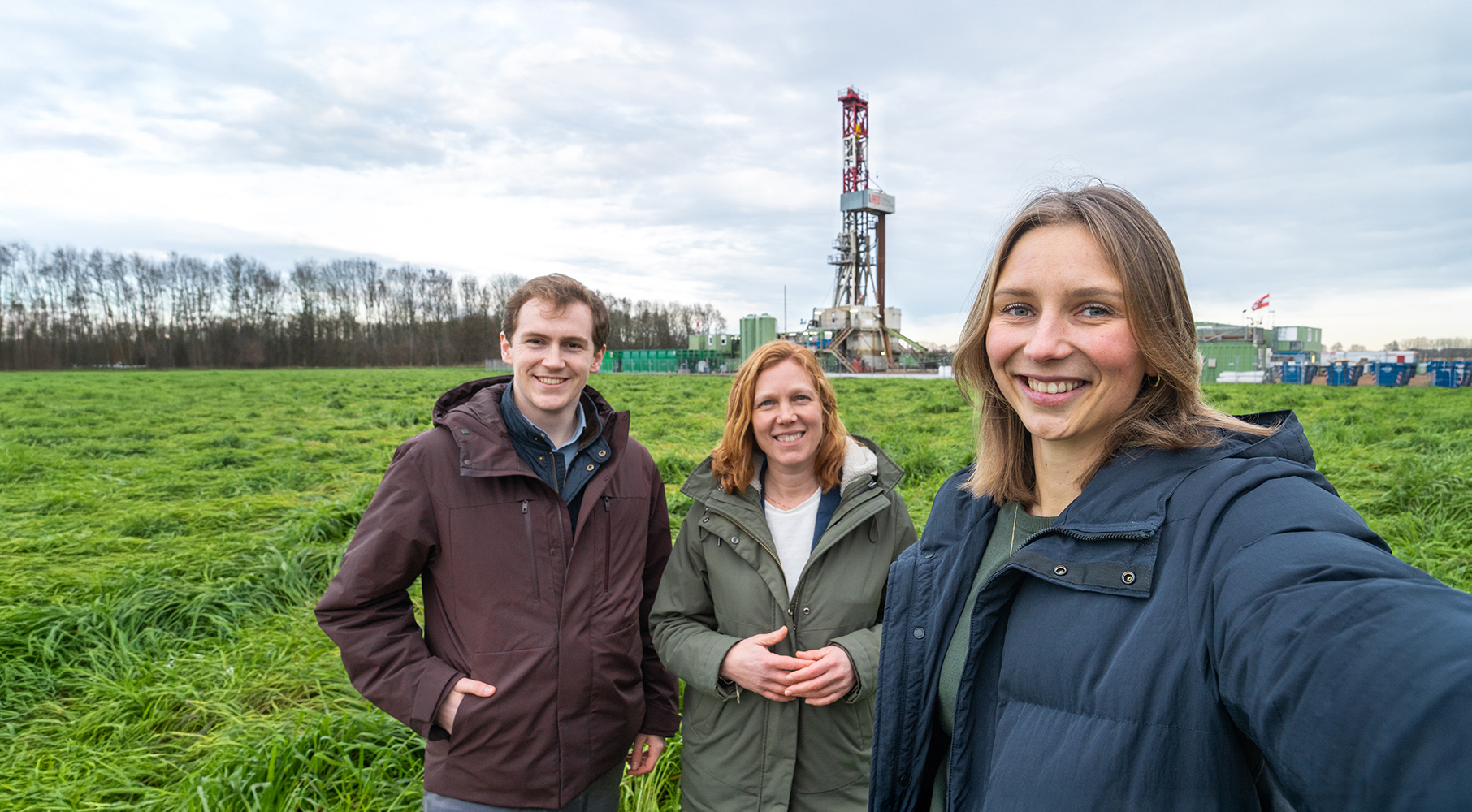 Drie EBN-medewerkers staan op een weiland met een boortoren en installaties op de achtergrond.