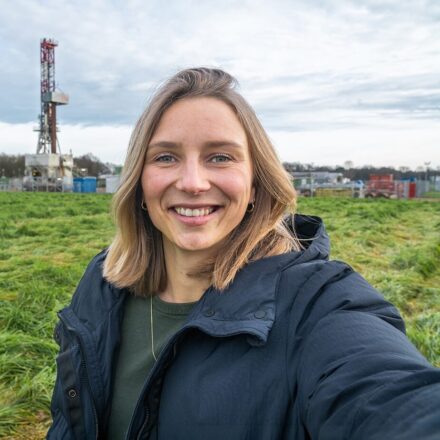 EBN-medewerker staat op een groen weiland met op de achtergrond een boortoren voor onderzoek naar aardwarmte.