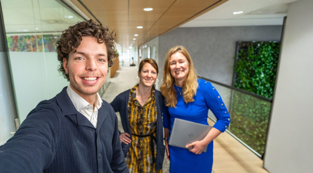 Drie EBN-medewerkers maken een selfie in een gang van het EBN-kantoor. Het kantoor is modern en licht met glazen wanden en een groene plantwand.