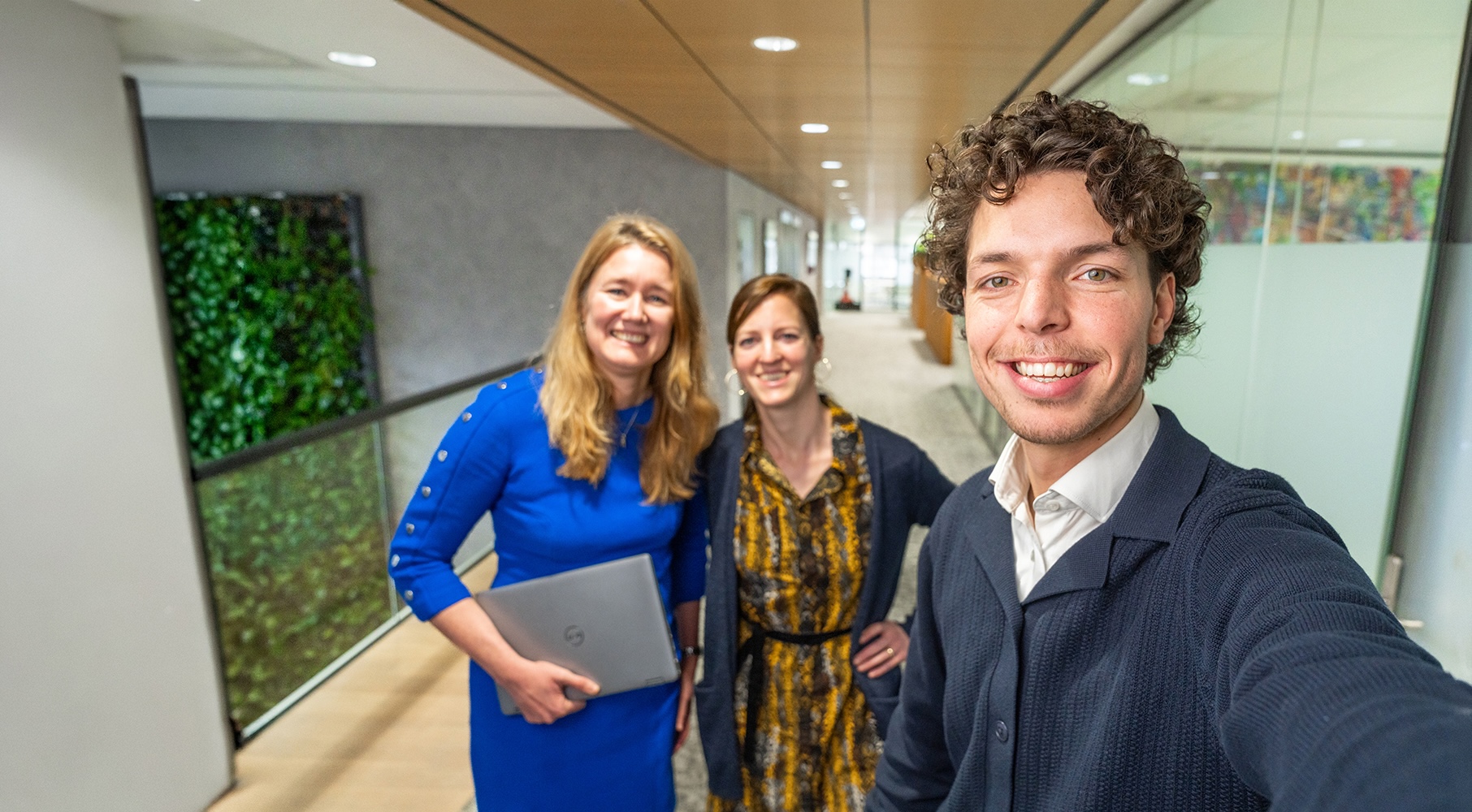 Drie EBN-medewerkers maken een selfie in een gang van het EBN-kantoor. Het kantoor is modern en licht met glazen wanden en een groene plantwand.