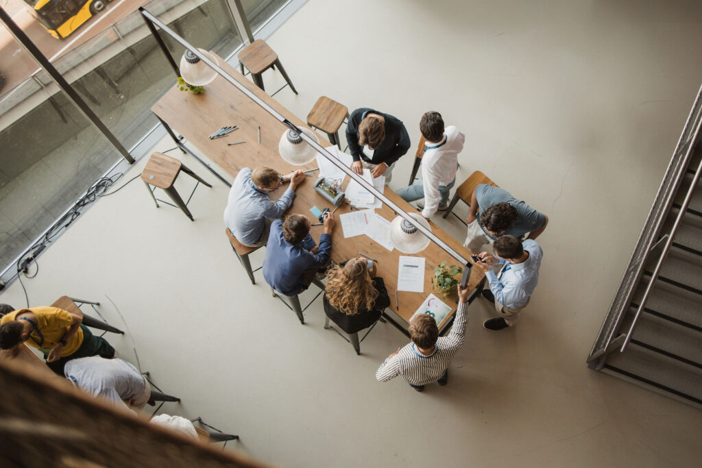 Van bovenaf gefotografeerde groep mensen rond een lange tafel in een lichte ruimte. Op de tafel liggen documenten, pennen en een technisch model terwijl deelnemers samenwerken en overleggen.