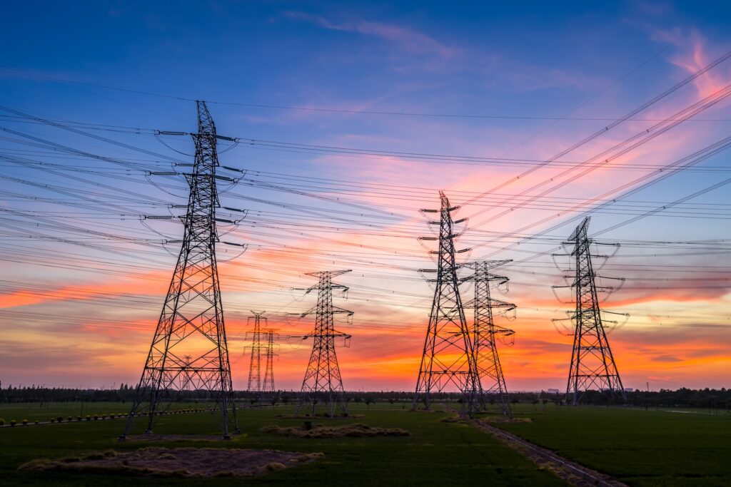 Hoogspanningsmasten en bovengrondse elektriciteitsleidingen in een groen landschap tijdens zonsondergang, met een kleurrijke lucht in blauw-, roze- en oranjetinten.